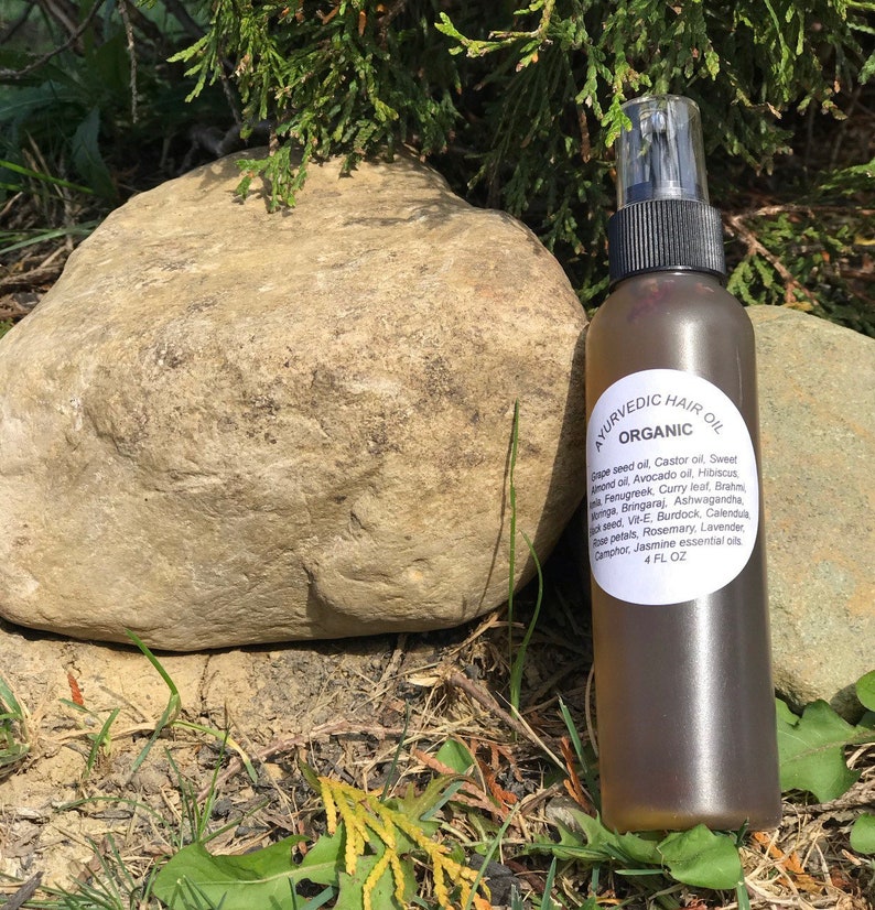 May include: A brown glass bottle with a spray nozzle, labeled "AYURVEDIC HAIR OIL ORGANIC" and listing ingredients including grape seed oil, castor oil, sweet almond oil, avocado oil, hibiscus, fenugreek, curry leaf, brahmi, amla, bringaraj, ashwagandha, black seed, vit-e, burdock, calendula, rose petals, rosemary, lavender, camphor, jasmine essential oils. The bottle is sitting on a rock and green grass.