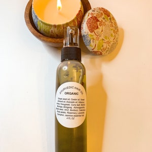 May include: A clear glass bottle with a brown spray top filled with a yellow liquid. The bottle label reads "Ayurvedic Hair Oil Organic" and lists a variety of ingredients including grape seed oil, castor oil, sweet almond oil, avocado oil, hibiscus, fenugreek, curry leaf, brahmi, moringa, bringaraj, ashwagandha, black seed, vitamin E, burdock, calendula, rose petals, rosemary, lavender, camphor, and jasmine essential oil. The bottle is labeled 4 fl oz.