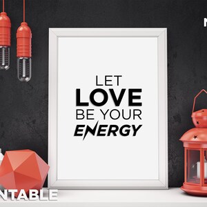Puede incluir: Una impresi&oacute;n enmarcada en blanco con el texto "Let Love Be Your Energy" en letras negras. La impresi&oacute;n est&aacute; sobre una mesa con una linterna roja, dos velas y una forma geom&eacute;trica roja.