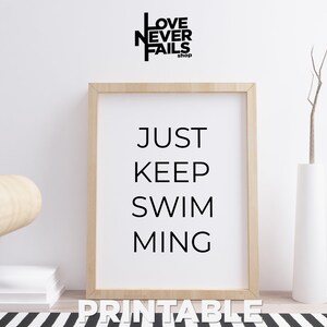 Puede incluir: Una impresión de arte de pared imprimible con fondo blanco y texto negro que dice "JUST KEEP SWIMMING". La impresión está enmarcada en un marco de madera claro y está sobre una alfombra de rayas blancas y negras.