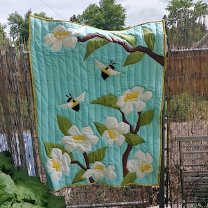Op de afbeelding: Een turquoise blauwe, gestikte wandhanger met een ontwerp van witte bloemen en gele en zwarte bijen. De bloemen zijn gemaakt van stof en zijn in een tak-achtig patroon gerangschikt.