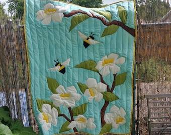 May include: A turquoise blue quilted wall hanging with a design of white flowers and yellow and black bees. The flowers are made of fabric and are arranged in a branch-like pattern.