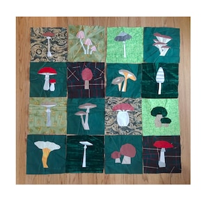 May include: A patchwork quilt with sixteen squares, each displaying a different mushroom design. The mushrooms are made from fabrics in red, green, white, and brown. The quilt is set against a wooden background, creating a rustic aesthetic.