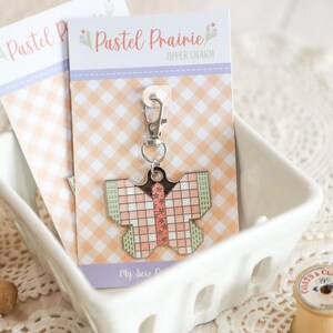 May include: A butterfly-shaped zipper charm with a pink and green grid pattern, attached to a silver clasp. The charm is displayed on a card with the text "Pastel Prairie Zipper Charm" and a gingham background, inside a white ceramic dish.