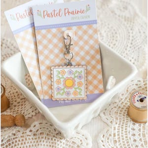 May include: A square floral zipper charm with a scalloped edge, featuring a central yellow flower surrounded by pastel blooms. The charm hangs from a silver clasp attached to a card with the text "Pastel Prairie". The background is a peach and white gingham pattern.
