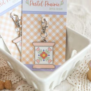 May include: A pastel-colored zipper charm with a floral design, featuring a spool shape and a silver-tone clasp. The charm is displayed on a card with a gingham pattern and the text "Pastel Prairie".