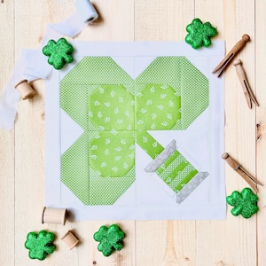 May include: A green shamrock-shaped quilt block with a white background. The shamrock is made of different green fabrics with white polka dots and a white floral pattern. A spool of thread is in the center of the shamrock.