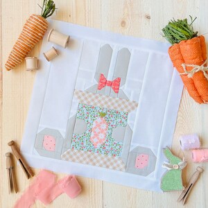 May include: A white fabric panel with a patchwork design featuring a bunny rabbit with a pink bow. The bunny is holding a small basket with a floral pattern and a pink and white checkered border. The panel is surrounded by a white border.