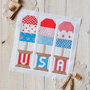 May include: A white fabric panel with three red, white, and blue popsicle-shaped fabric blocks. Each block has a different pattern and is stitched with the words "Stitched" and "Stitched" on the bottom. The words "USA" are stitched in red, white, and blue on the left block, "USA" is stitched in blue, white, and blue on the middle block, and "USA" is stitched in red, white, and blue on the right block.