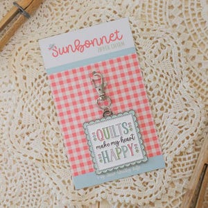 May include: A Sunbonnet zipper charm with a metal clasp. The charm features a square design with the words "QUILTS make my heart HAPPY" in a whimsical font, surrounded by hearts. The background is a pink and white gingham pattern.