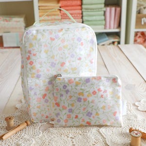 May include: A white travel bag and matching pouch, both adorned with a delicate floral pattern in pastel colors. The bag has a handle and a zipper closure. The pouch has a zipper closure. The floral design includes orange, yellow, purple, and pink flowers.