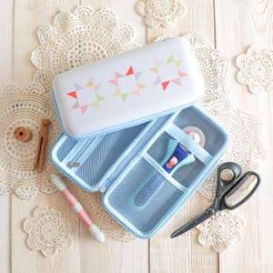 May include: A light blue sewing kit with a white lid featuring colorful star designs. Inside, a rotary cutter, scissors, and other sewing tools are visible. The kit is open, revealing mesh pockets and a zipper closure.