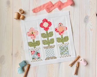 Könnte beinhalten: Ein weißes Stoffpanel mit drei Blumen-Patches, jedes mit einem anderen Blumendesign und einem Topf mit einem anderen Muster. Das Panel befindet sich auf einem weißen Holzgrund.