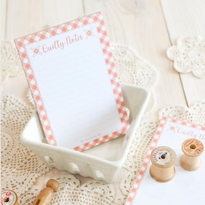 May include: A white notepad with a peach gingham border and the text "Quilty Notes" sits in a white ceramic dish. The notepad is lined and has floral accents. Wooden spools and a wooden clothespin are also visible.