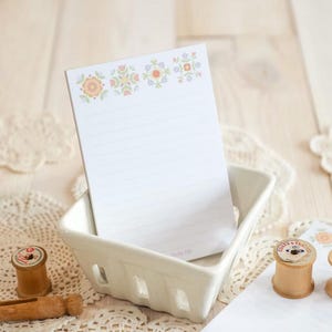 May include: A white lined notepad with a floral design in peach, green, and lavender sits in a white ceramic basket. The notepad has the text "Quilty Life" at the bottom. Wooden spools of thread and lace doilies are in the background.