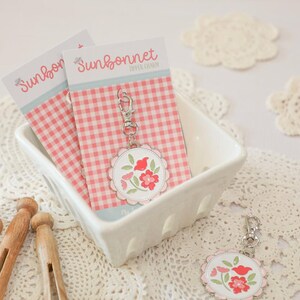 May include: Keychains with floral designs and the text "Sunbonnet" on a pink gingham background. The keychains are in a white ceramic dish, with wooden clothespins and a spool of thread nearby. The background includes white lace doilies.