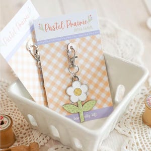 May include: A white ceramic dish holds two flower-shaped zipper charms. Each charm has a white flower with a yellow center and green leaves. The charms are attached to cards with the text "Pastel Prairie" and "Zipper Charm".