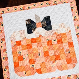 Pu&ograve; includere: Un quadrato di tessuto bianco trapuntato con un fiocco bianco e nero sopra una zucca patchwork realizzata con quadrati di tessuto arancione e giallo. Il tessuto &egrave; decorato con un motivo di gatto nero e zucca.