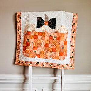 Pu&ograve; includere: Un trapunta bianca con un design a forma di zucca arancione e nera. La trapunta &egrave; appesa a una scala bianca contro un muro beige.