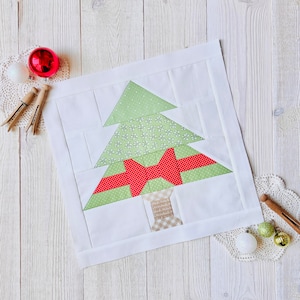 May include: A white fabric Christmas tree quilt block with a green and red bow. The tree is made of patchwork fabric with a green and white floral pattern. The block is on a white wooden surface.