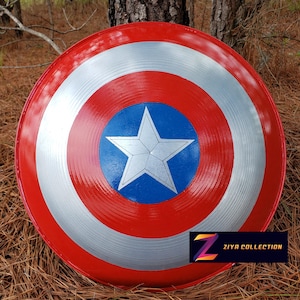 May include: A red, white, and blue shield with a star in the center, reminiscent of a superhero's emblem. The shield has a glossy finish and is displayed outdoors. The text "ZIYA COLLECTION" is visible on the lower right.
