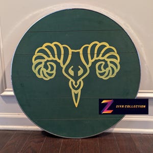 May include: A round, wooden wall decor piece with a dark green background. A gold-colored ram's head design is centered on the piece. The text "ZIYA COLLECTION" is in a rectangular box in the lower right corner.