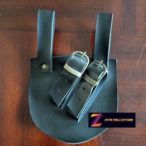 Puede incluir: Bolsa de cuero negro con dos correas y hebillas metálicas. La bolsa tiene una base redondeada y dos correas verticales para la fijación. El texto "Ziya Collection" es visible en la esquina inferior derecha.