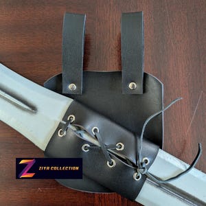 Puede incluir: Una vaina de espada de cuero negro con remaches plateados y un cierre con cordones, diseñada para sujetar una espada. La vaina tiene dos correas para sujetarla y presenta el logotipo "Ziya Collection".
