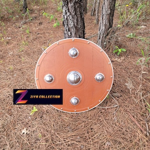 May include: A round, brown shield with a white border and four silver-colored studs. The shield has a central, larger stud. The text "ZIYA COLLECTION" is displayed on a rectangular label in the lower left corner. The shield is outdoors.