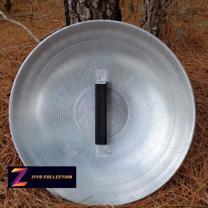 May include: A large, circular, silver-colored metal shield with a black handle. The shield has a ridged pattern radiating from the center. The text "ZIYA COLLECTION" is displayed on a black rectangle with a colorful "Z" logo.