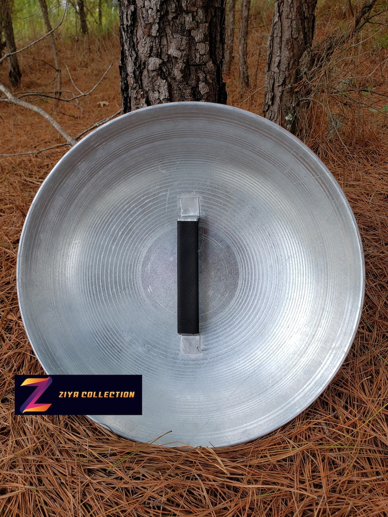 May include: A large, circular, silver-colored metal shield with a black handle. The shield has a ridged pattern radiating from the center. The text "ZIYA COLLECTION" is displayed on a black rectangle with a colorful "Z" logo.