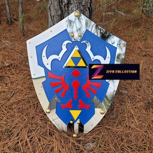 May include: A blue, white, and silver shield with gold accents. The shield features a red emblem and a yellow triangle. The text "ZIYA COLLECTION" is displayed on the shield. The shield is resting on a bed of pine needles.