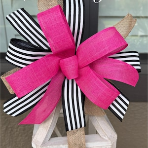 May include: A decorative bow with a bright pink centre, black and white striped ribbons, and natural burlap. The bow is attached to a white wooden lantern. The text "Lorell's Designs" is visible.
