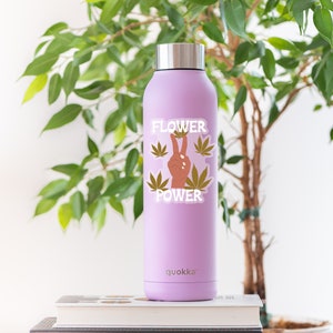 Puede incluir: Una botella de agua de acero inoxidable de color morado con una pegatina que dice "Flower Power" y que presenta un signo de paz y hojas de marihuana. La botella est&aacute; sobre una pila de libros.