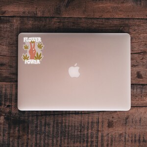 Puede incluir: Un ordenador port&aacute;til plateado con un logotipo de Apple blanco en la tapa. Una pegatina con el texto "Flower Power" y un signo de paz con hojas verdes est&aacute; en el lado izquierdo del port&aacute;til.