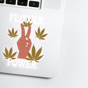 Puede incluir: Una pegatina con un signo de paz formado por una mano con dos dedos levantados. La mano es marr&oacute;n y los dedos apuntan hacia arriba. La pegatina muestra las palabras "FLOWER POWER" en texto blanco, rodeadas de cuatro hojas verdes de marihuana.