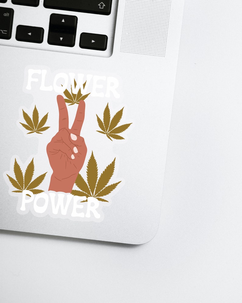 Puede incluir: Una pegatina con un signo de paz formado por una mano con dos dedos levantados. La mano es marr&oacute;n y los dedos apuntan hacia arriba. La pegatina muestra las palabras "FLOWER POWER" en texto blanco, rodeadas de cuatro hojas verdes de marihuana.