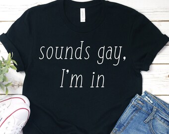 May include: Black t-shirt with the text "sounds gay, I'm in" in white. The shirt is paired with denim shorts, white trainers, and a small green plant. The items are arranged on a light wood-grain surface.