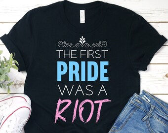 Könnte beinhalten: Schwarzes T-Shirt mit dem Text "The First Pride Was A Riot" in blauer und rosa Schrift.
