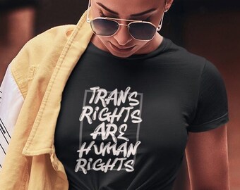 Könnte beinhalten: Ein schwarzes T-Shirt mit weißem Text, der "Trans Rights Are Human Rights" in einem Kasten lautet. Das Shirt wird von einer Person mit einer gelben Jacke und einer blauen Jeans getragen.