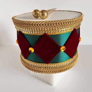 May include: A decorative drum with a white top and a green body adorned with red diamond shapes and gold trim. The drum features gold drumsticks and is embellished with gold accents and jewels.