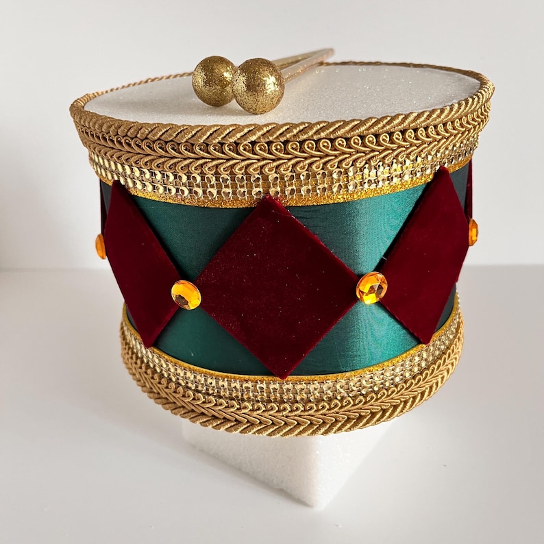 May include: A decorative drum with a white top and a green body adorned with red diamond shapes and gold trim. The drum features gold drumsticks and is embellished with gold accents and jewels.