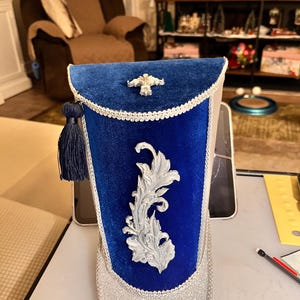 May include: A decorative blue and silver hat with a silver floral design and a blue tassel. The hat has a blue velvet top with a silver trim and a silver glitter base. A silver fleur-de-lis is on the top.