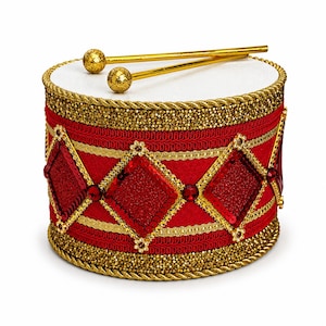 May include: A cylindrical drum featuring a vibrant red fabric body decorated with gold braided trim, sparkling red diamond-shaped sequin panels, and small red gems. The top surface is white, and two gold-colored drumsticks with glittery ball ends rest on top.