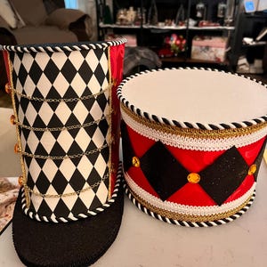 May include: Two decorative drum-shaped containers. One is tall with a black and white diamond pattern, gold chain accents, and a black base. The other is shorter, with red and black diamond shapes, gold trim, and a white top. Both have rope trim.