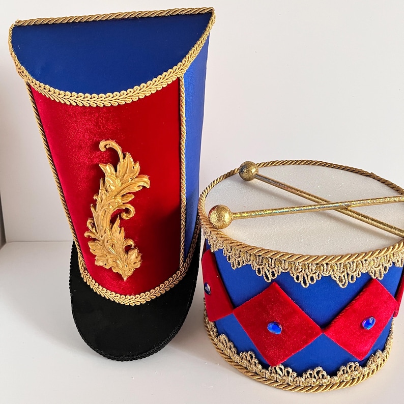 May include: A decorative toy drum and tall hat in a patriotic theme. The drum is blue with red diamond accents and gold trim. The hat is blue and red with gold trim and a gold leaf design. The drumsticks have gold ball ends.