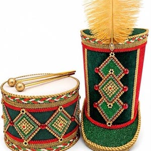 May include: A green and red drum with two gold drumsticks and a matching tall hat featuring a gold feather plume. Both items are decorated with gold braided trim, diamond-shaped patterns, and red beads. The text Mercier Handcrafts is visible.