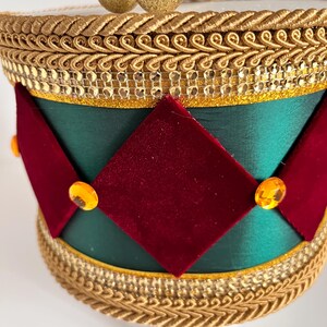 May include: A decorative hat with a green satin base, adorned with red velvet diamond shapes and gold trim. The hat features gold embellishments and orange-colored jewels, creating a festive and elegant design.