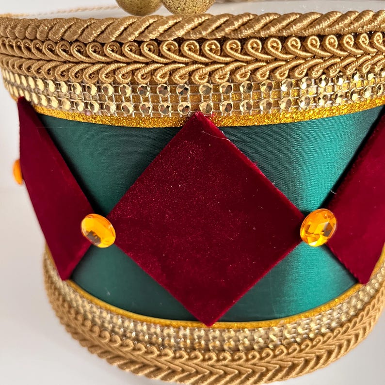 May include: A decorative hat with a green satin base, adorned with red velvet diamond shapes and gold trim. The hat features gold embellishments and orange-colored jewels, creating a festive and elegant design.