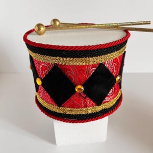 May include: A decorative drum with a white top, red and black patterned sides, and gold accents. The drum features black diamond shapes, gold trim, and red rope detailing. Two gold drumsticks rest on top. The drum is likely a decorative item.